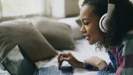Close-up of african american girl listen to music while watching photos online on laptop lying on...