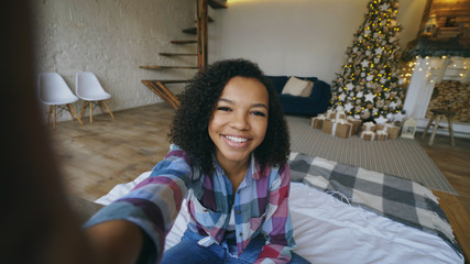 Funny mixed race girl taking selfie pictures on smartphone camera at home near Christmas tree