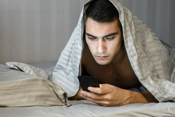 Closeup portrait sleepy young man messaging on smart phone in bed at home, suffering from insomnia, modern society's problems. the concept-social networking, Internet, communication and people