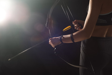 Sporty young woman with bow and arrow for practicing archery on dark background