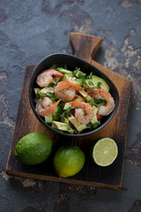Bowl of avocado salad with shrimps and cucumber on a wooden serving board, brown stone background, vertical shot