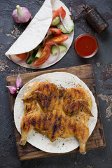 Roasted chicken tabaka served with tortilla bread and fresh vegetables. View from above on a brown stone background