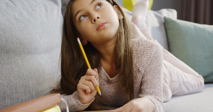Pretty teen girl lying on the grey couch with a pencil in a hand and thinkig what to write in the notebook. At home. Inside