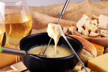 Gourmet Swiss fondue dinner on a winter evening with assorted cheeses on a board alongside a heated pot of cheese fondue with two forks dipping bread and white wine behind in a tavern or restaurant