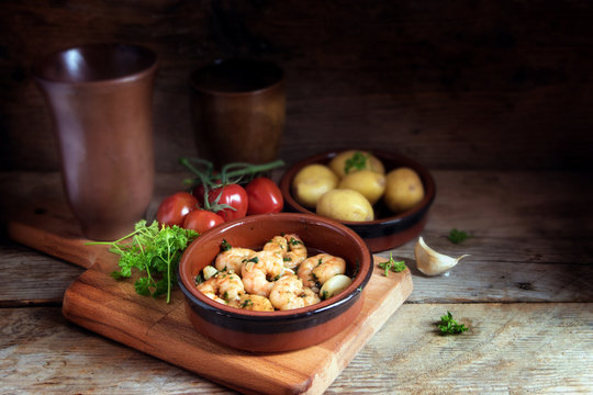 Tapas bowl with shrimps or prawns in garlic olive oil, potatoes, tomatoes and herbs on a rustic wooden table, spanish appetizer, dark background with cop space