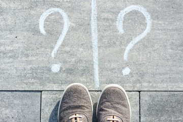 Two question marks are handwritten on an asphalt road with sneakers