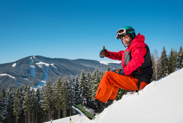 Shot of a happy snowboarder sitting, relaxing on top of the mountain, showing thumbs up to the...
