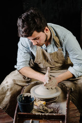 pottery, workshop, ceramics art concept - man working on potter's wheel with raw clay with hands, a male brunette sculpt a utensils near wooden table with tools, master in apron and a shirt
