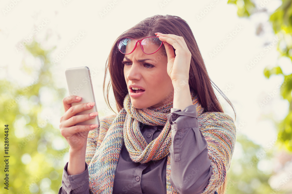 Wall mural annoyed upset woman in glasses looking at her smart phone with frustration while walking on a street