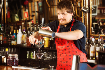 Bartender is pouring alcohol drink using strainer and shaker at bar background.