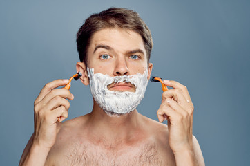 Man with a beard on a gray background in shaving foam holds razors, portrait