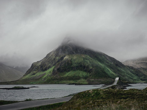 Gro&szlig;er markanter Berg steht in nebelig wolkiger Landschaft . Eine Stra&szlig;e mit Br&uuml;cke davor l&auml;uft auf Ihn zu umgeben von Wasser