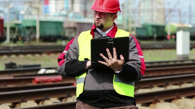 Railway worker with tablet pc at train terminal. Railwayman in uniform and red hard hat works with tablet computer. Railway employee makes notes in his tablet pc