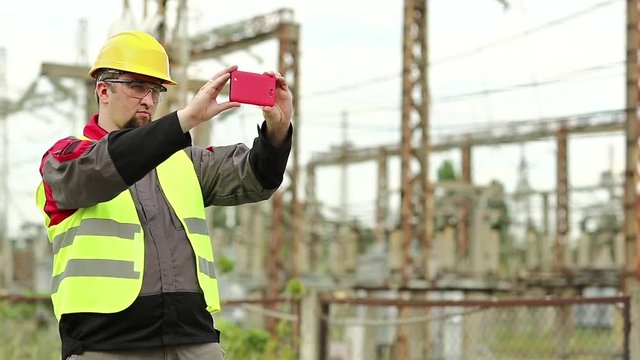 Worker records videos on smartphone. Powerman in yellow hard hat with smartphone at electric power plant