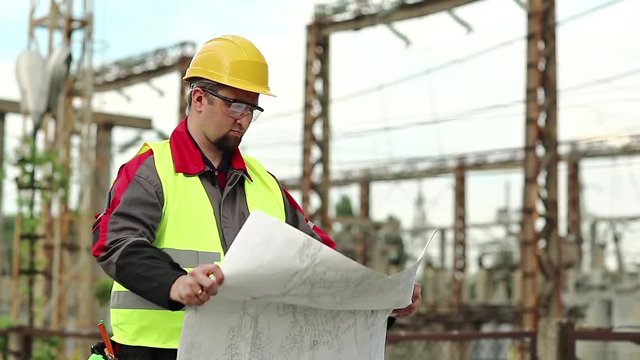 Worker with scheme of works on power station. Power man with construction documentation at power plant. Master builder in hard hat with design drawings