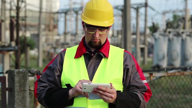Electrician at power plant uses smartphone. Worker uses cell phone at heat electric power station near outdoor switchgear