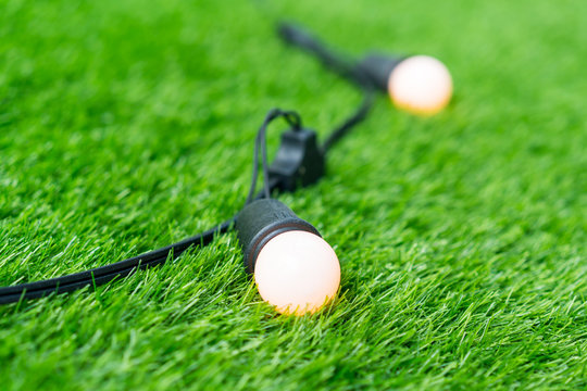 Closed Up Orange Illuminated Lightbulb With Black Nylon Braided Cover Cord And Extension On Green Grass Background