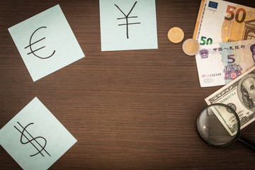 Banknotes and coins lying on notepad, stickers with currency signs and Magnifier on wooden table. Accounting and taxes. Dollar, Euro, Yuan Renminbi money. Budget ca