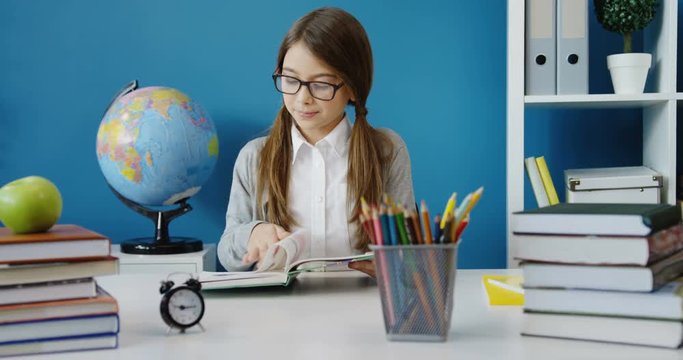 Lovely teenager girl in glasses reading a book and scrolling pages while sitting at the table with different stuff in the office ot classroom. Indoor