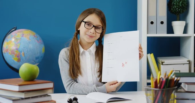 Portrait shot of the lovely teeen girl in glasses showing a paper sheet with her test and grade A+ on it while sitting at the working desk. Indoor
