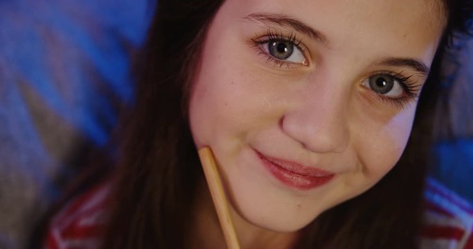 Close up of the pretty smiled teen girl's face with pencil. Girl thinking about something and smiling. At night. At home. Indoor. Portrait