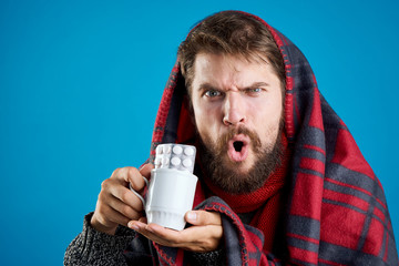 a man with a beard on a blue background in a blanket holding a cup of pills, illness, flu, sick, runny nose