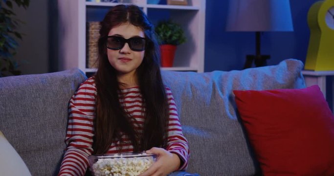 Funny teenager girl in a 3D glasses sitting on the grey sofa with pillows, eating popcorn and watching a comedy in the living room at late night. Inside