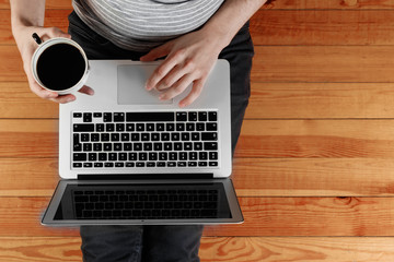 A laptop and a cup of coffee in the hands of a man sitting on a wooden floor. View from above, copy space.