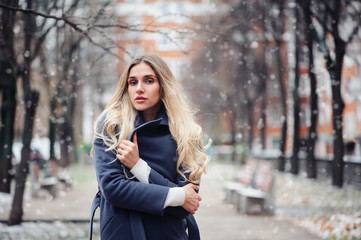 winter portrait of young woman walking on city streets in snowy day, wearing warm grey coat
