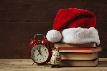 Red alarm clock and statue of Santa Claus. On the books Santa's hat, on a wooden background