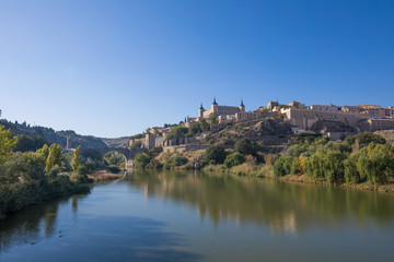 Fototapeta na wymiar Scenery of Toledo city, in Spain, Europe. Green water river Tagus, Alcantara arch bridge, landmark and monument from ancient Roman age, and alcazar building 