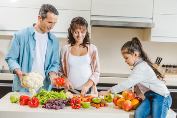 cheerful family with pregnant mother slicing vegetables and fruits at kitchen