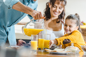 happy family at table, man pouring juice in glasses to woman and kid at kitchen