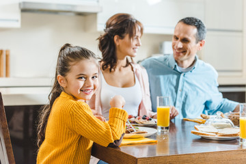 happy family sitting at table with pancakes and juice, little kid smiling and looking at camera