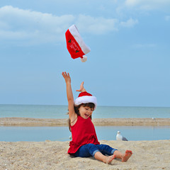 Cute little girl celebrating Christmas and New Year holidays on the beach, throwing up Santa hat
