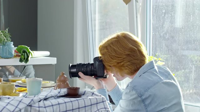 PAN of young redhead man kneeling and taking still life pictures of food on digital camera
