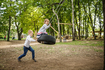 The brother rolls the younger sister on a tire swing. Children playing outdoors in summer.