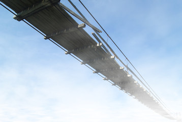 view from below on an old wooden suspension bridge against a sky hiding in clouds in perspective