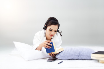 lady in white shirt is sitting on blady in white shirt is read book and listen music by headphone,on bed,blurry light around.ed