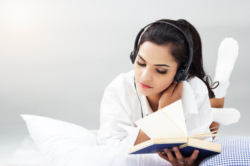 lady in white shirt is sitting on lady in white shirt is read book and listen music by headphone,on bed,blurry light around.bed