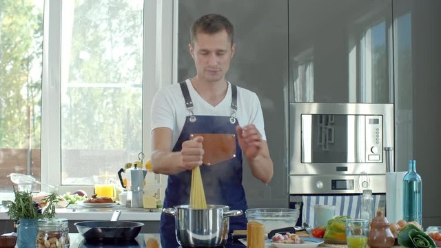 Medium shot of male cook in apron putting spaghetti into pot with boiling water and salting it while talking to camera in modern kitchen
