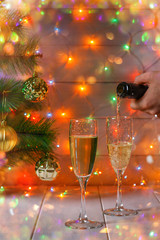 Men's hands pour champagne from a bottle in glasses against the background of a New Year tree and garlands.