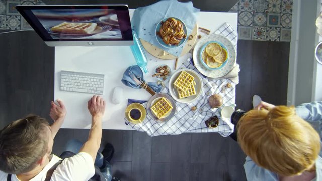 Top view of young male photographer taking still life pictures of breakfast food lying on plates on checkered tablecloth while his assistant looking at photos on computer screen