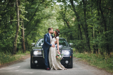 Newlyweds near the black wedding car stand on the road in the summer forest.