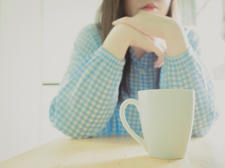 white cup of hot coffee for drink in morning time with soft focus (asian girl in blue pajamas at her bedroom with both hand support her chin) background