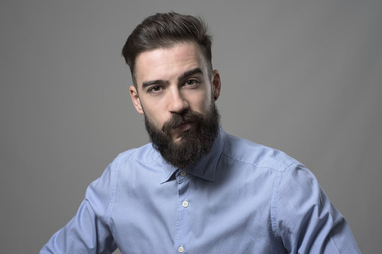 Skeptical suspicious young bearded stylish businessman looking at camera with one eyebrow raised against gray studio background. 