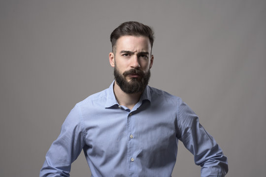 Young Serious Upset Bearded Business Man With Arms On Hips And Intense Look At Camera Against Gray Studio Background. 