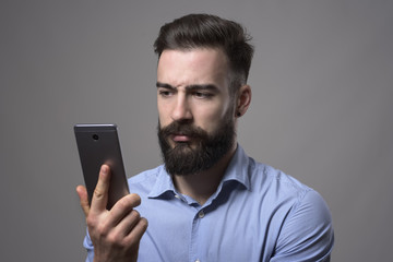 Serious dissatisfied young bearded man holding and watching smartphone against gray studio background. 