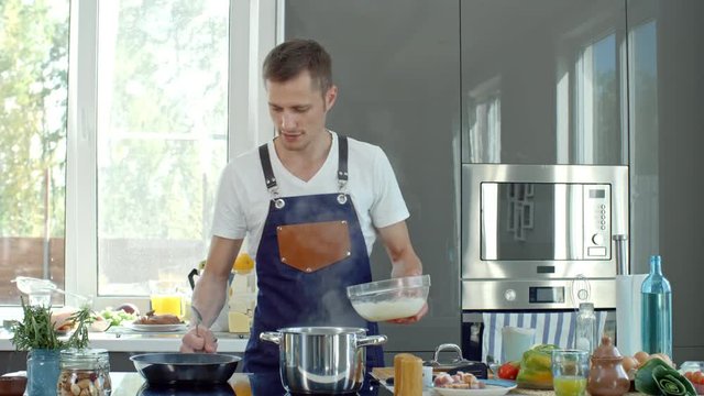 Medium shot of cooking show presenter in apron standing at kitchen countertop and adding cream sauce into hot pan, then stirring with spoon while explaining recipe before camera