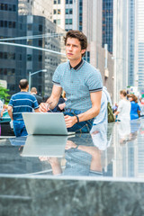 European college student study in New York. Wearing blue pattered shirt, holding glasses, a young handsome guy working on laptop computer on street, looking up, thinking. Many people on background..